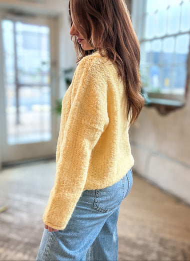 A woman wearing a fuzzy, textured pale yellow long-sleeve sweater and light blue jeans, standing in a bright, modern indoor space with large windows.