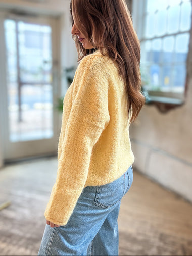 A woman wearing a fuzzy, textured pale yellow long-sleeve sweater and light blue jeans, standing in a bright, modern indoor space with large windows.