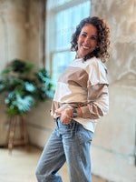 A woman with curly brown hair stands in a bright, modern loft space wearing a color-blocked long-sleeve top with beige and white panels, paired with light-wash blue jeans.