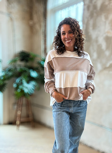 A woman with curly brown hair smiling while wearing a beige and white striped long-sleeve top and blue jeans, standing in a bright, modern indoor space with concrete walls and a wooden floor.