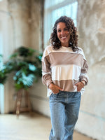 A woman with curly brown hair smiling while wearing a beige and white striped long-sleeve top and blue jeans, standing in a bright, modern indoor space with concrete walls and a wooden floor.