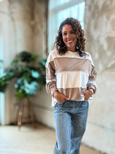 A woman with curly brown hair smiling while wearing a beige and white striped long-sleeve top and blue jeans, standing in a bright, modern indoor space with concrete walls and a wooden floor.