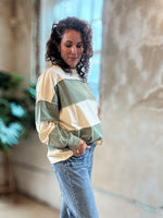 A woman with curly dark hair stands in a loft-style room wearing a color-blocked long-sleeve top with beige, sage green, and cream panels, paired with wide-leg blue jeans and brown boots.