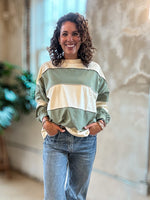 A woman with curly brown hair stands in a loft-style room wearing a sage green and cream color-blocked sweatshirt and light-wash blue jeans.