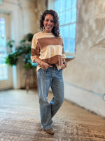 A woman stands in a bright, rustic indoor space wearing a color-blocked long-sleeve top with beige, brown, and cream horizontal stripes, paired with light-wash blue jeans and brown shoes.